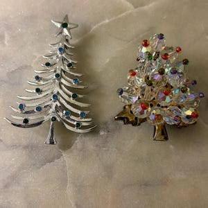 May include: Two Christmas tree brooches. One is a silver-tone metal tree with a star topper and small blue and green rhinestones. The other is a gold-tone metal tree adorned with clear faceted crystal beads featuring small colorful accents.
