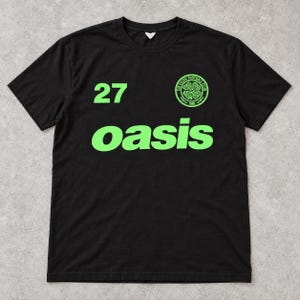 May include: A black short-sleeved t-shirt featuring the word oasis in bright green lettering across the chest. The number 27 is printed in green on the upper left, and a circular green and black Celtic Football Club logo is on the upper right.