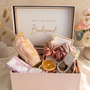 Peut inclure: Une boîte cadeau rose contenant un gobelet en verre avec une paille, un carnet à motifs violets, une tranche d'orange séchée, un bracelet de perles et deux chouchous en satin. Le couvercle porte l'inscription Will You Be My Bridesmaid en doré.