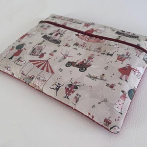 Può includere: Una custodia rettangolare imbottita con una stampa stravagante con illustrazioni a tema circense in rosso, rosa e beige. La custodia ha una cerniera e un bordo bordeaux. Il design include figure, veicoli ed elementi festivi.