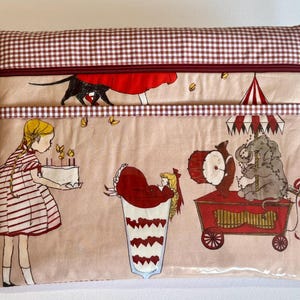 Puede incluir: Bolsa rectangular con un estampado caprichoso con temática de circo. El diseño presenta ilustraciones de una niña con un pastel, un elefante, un oso y un payaso. La parte superior y la cremallera están adornadas con un estampado a cuadros rojos y blancos.