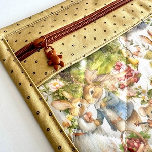 Può includere: Un astuccio trapuntato con una stampa di Peter Rabbit. Presenta un bordo dorato a pois e una cerniera marrone con un ciondolo a forma di orsetto. Il tessuto interno mostra una scena affascinante con conigli, fragole e fiori.