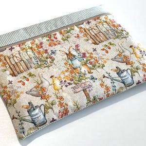 Könnte beinhalten: Eine rechteckige Stofftasche mit Reißverschluss auf beigem Grund, bedruckt mit Hasen, Gänsen, Gießkannen und Blumen. Der obere Teil ist mit einem hellgrün-weißen Vichy-Muster versehen. Ein metallener blumenförmiger Anhänger ist am Reißverschluss befestigt.