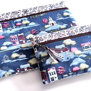 May include: Two rectangular zippered pouches in different sizes, featuring a blue fabric with a printed pattern of houses, trees, and bushes. The top edge of each pouch has a floral print border, and the zippers include decorative ribbon pulls.