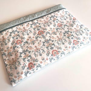 Puede incluir: Una bolsa rectangular con cremallera y un estampado floral en tonos melocotón, azul y crema. Tiene un borde acolchado en color gris claro. La cremallera es negra.