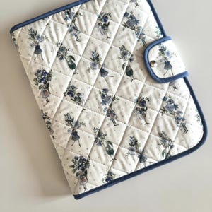 Può includere: Una custodia rettangolare in tessuto trapuntato con base bianca e un motivo ripetuto di bouquet di fiori blu e piccoli punti grigi. La custodia presenta un bordo in tessuto blu lungo i lati e una chiusura a patta con bottone a pressione.