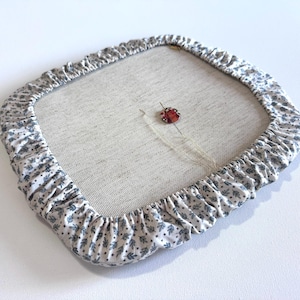 Puede incluir: Bastidor de bordado cuadrado con un centro de tela de lino beige claro. La tela está rodeada por un borde con volantes con un fondo blanco y un estampado floral azul. Un alfiler de mariquita rojo y negro está sujeto a la tela con aguja e hilo.