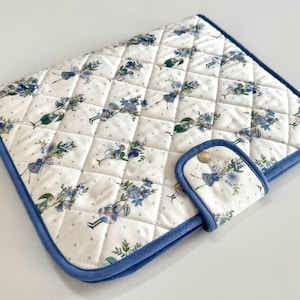 Könnte beinhalten: Eine gesteppte Stoffhülle mit weißem Hintergrund und einem wiederkehrenden Muster aus blauen Blumensträußen und kleinen hellblauen Punkten. Die Hülle hat eine blaue Stoffeinfassung, eine rechteckige Form und eine Lasche mit Metalldruckknopf.