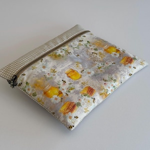 Pode incluir: Uma bolsa retangular com zíper com um padrão de abelhas e potes de mel. O tecido principal apresenta potes de mel amarelos, abelhas e flores brancas em um fundo cinza. A borda superior tem um padrão xadrez bege e branco.