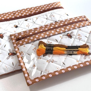 Può includere: Due pochette rettangolari trapuntate con cerniera, realizzate in tessuto bianco con un motivo di api e piccoli fiori. Le pochette presentano un bordo marrone con stampa floreale bianca e una finestra in plastica trasparente. Tre matasse di filo sopra.