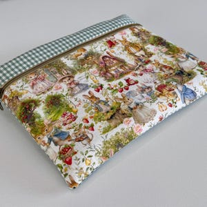 Puede incluir: Una bolsa rectangular con cremallera que presenta un estampado caprichoso de conejos, flores y fresas sobre un fondo blanco. El borde superior está adornado con un patrón de cuadros vichy verde y blanco. La cremallera es de color bronce.