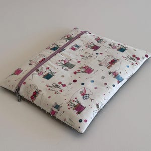 May include: A rectangular, off-white sewing machine case with a zipper closure. The case is patterned with illustrations of sewing spools, flowers, and sewing tools in pink, blue, and green. The zipper is lavender.