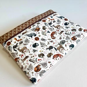 Puede incluir: Una bolsa rectangular con cremallera con un fondo blanco y un patrón repetido de animales del bosque, helechos y setas en tonos marrón, gris y naranja. El borde superior y la cremallera son marrones. La bolsa es de tela.