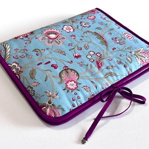 Könnte beinhalten: Hellblau gesteppte Notizbuchhülle mit einem Blumenmuster in Rosa, Braun und Grün. Die Hülle hat eine dunkelviolette Einfassung und eine passende Schleife. Das florale Design zeigt große Blüten und Ranken.