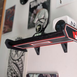 May include: A black wall-mounted shelf featuring a racing-inspired spoiler design with red and white trim accents. The shelf is supported by two black brackets and holds a small white digital clock with a circular display on its top surface.