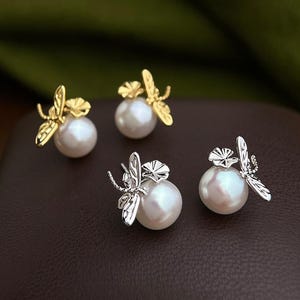 Op de afbeelding: Twee paar oorknopjes met witte bolvormige parels en metalen libellemotieven. Eén paar is afgewerkt in een glanzende goudtint, terwijl het andere paar is afgewerkt in een gepolijste zilverkleur, elk met gedetailleerde vleugels.