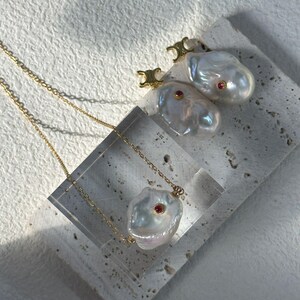 May include: A gold chain necklace with a large, irregular pearl pendant and a small red gemstone. Two matching pearl earrings with gold accents and red gemstones are also displayed. The jewellery is set against a neutral background.