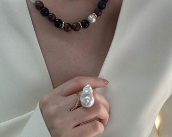 May include: A necklace with large, dark brown, black, and white beads, interspersed with gold accents. A large, baroque pearl ring with a gold band and small accent stones is also visible.