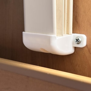 Pode incluir: Um suporte de plástico branco montado numa superfície de madeira com um parafuso de metal prateado. O suporte segura um componente vertical de plástico branco com lâminas internas visíveis, fixando-o contra o painel de madeira castanha.