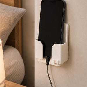 May include: A white plastic wall-mounted phone holder with a rectangular pocket design. The holder is attached to a vertical surface and contains a black smartphone connected to a charging cable that extends downward from the bottom of the device.