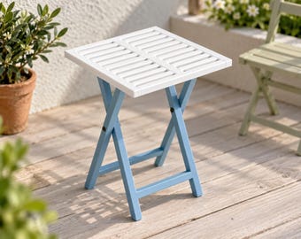 Puede incluir: Una mesa plegable cuadrada con una superficie de listones blancos y una estructura de color azul claro. La mesa presenta un diseño de patas en forma de X con herrajes negros que conectan los componentes del marco.