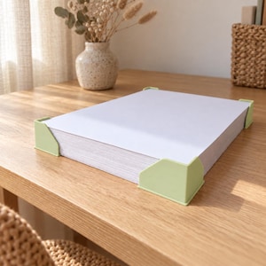 May include: A stack of white paper sheets secured at the corners with light green plastic corner protectors. The protectors are smooth and angular, designed to hold the paper stack together firmly on a flat wooden surface.