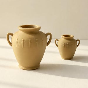 Puede incluir: Dos jarrones de color beige con acabado mate, cada uno con cuerpo redondeado, cuello estrecho y dos pequeñas asas laterales. El jarrón más grande está decorado con una serie de marcas verticales en relieve en forma de lanza en el hombro.