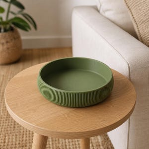 May include: A round, olive green tray with a smooth interior surface and a vertically ribbed exterior texture. The tray features a low, raised edge and a flat base, presenting a uniform matte finish across its entire circular structure.