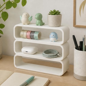 May include: A white three-tiered oval shelf organizer holding five rolls of patterned washi tape, a small white bird figurine, a green dinosaur figurine, a succulent in a speckled pot, a white earbud case, and a coiled charging cable.