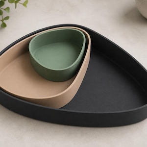 Peut inclure: Un ensemble de trois plateaux emboîtables de forme triangulaire arrondie irrégulière. Le plus grand plateau est noir mat, le moyen est beige terreux et le plus petit bol est vert sauge. Chaque pièce présente des bords lisses et incurvés.