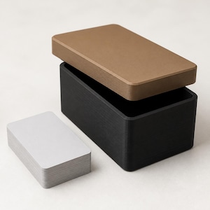 May include: A rectangular black storage box with a separate bronze-colored lid placed at an angle on top. Next to the box is a stack of plain, light gray rectangular cards with rounded corners, matching the dimensions of the box interior.