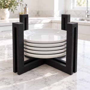 May include: A stack of five plain white ceramic plates held in place by a black wooden stand with four vertical pillars connected by a cross-shaped base. The stand features a textured finish and provides support for the plates.