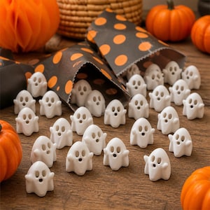 May include: A collection of small white ghost figurines with carved facial features, scattered on a wooden surface alongside orange pumpkins and black paper bags featuring an orange polka dot pattern.