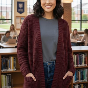 May include: A burgundy knit cardigan with two front pockets. The open-front sweater is worn over a gray t-shirt and blue jeans. The cardigan is made of a thick, textured knit material. The background includes bookshelves and a library setting.