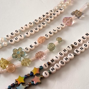 May include: Three beaded lanyards featuring white letter beads with black text. The first reads YES ITS SCARY BUT YOURE BRAVE, the second reads SEX SELLS, and the third reads I MIGHT OPEN I MIGHT KNOCK. They include various colorful glass beads.