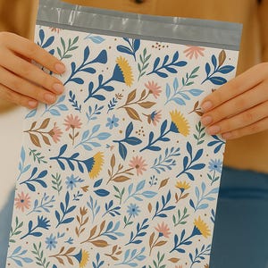 May include: A white floral patterned shipping envelope with a gray sealed top. The pattern includes blue, yellow, pink, and brown flowers and leaves. The envelope is held by two hands.