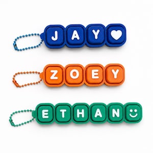 May include: Three rectangular keychains with rounded edges, each featuring a ball chain. The top blue keychain spells JAY with a heart, the middle orange one spells ZOEY, and the bottom green one spells ETHAN with a smiley face in white letters.