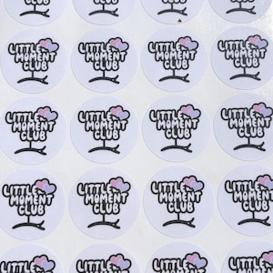 May include: A collection of white, circular stickers. Each sticker displays the words "LITTLE MOMENT CLUB" in black, accompanied by a cloud design in pink and purple hues and a small, curved line. The stickers are arranged in a grid.