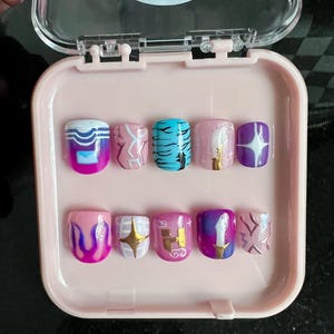 Puede incluir: Un juego de diez uñas postizas en un estuche rosa. Las uñas presentan varios diseños, incluyendo ondas, patrones de mármol, rayas de tigre, purpurina y estrellas. Los colores incluyen rosa, azul, morado, dorado y blanco.