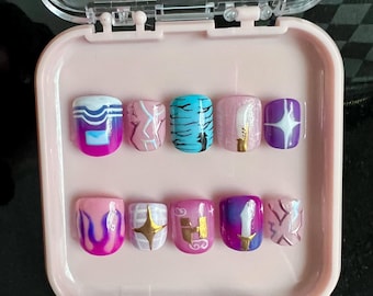Pode incluir: Um conjunto de dez unhas postiças numa caixa rosa. As unhas apresentam vários desenhos, incluindo ondas, padrões de mármore, listras de tigre, glitter e estrelas. As cores incluem rosa, azul, roxo, dourado e branco.