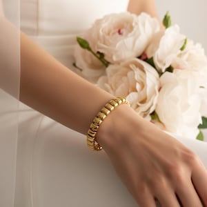 May include: A gold-colored bracelet with a ribbed design is worn on a wrist. The bracelet is the focal point, with a bouquet of white flowers in the background. The bracelet's design is made up of a series of rectangular links.