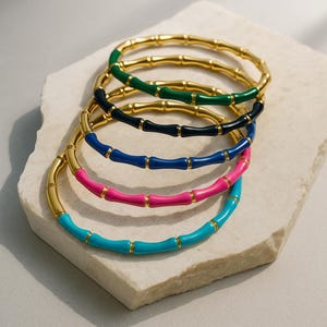 Può includere: Una collezione di braccialetti dorati in stile bambù con accenti smaltati colorati. I braccialetti presentano sezioni smaltate verdi, nere, blu, rosa e turchesi. I gioielli sono esposti su una superficie bianca strutturata.