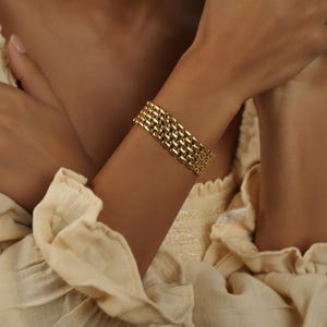 May include: A gold-coloured bracelet with a linked design. The bracelet is made of multiple rows of rectangular links, creating a textured accessory. The bracelet is worn on the wrist.
