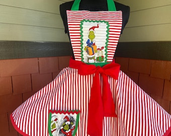 May include: A red and white striped apron featuring a central rectangular patch with a green character wearing a Santa hat and holding a gift. The apron has a large red waist tie and a pocket at the bottom with a matching green character graphic.