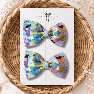 May include: Two hair bows attached to a white display card labeled Simply Knotty bows. The bows feature a colorful pattern of cartoon characters including a green one-eyed monster, a large blue furry creature, and a small child on a gradient background.