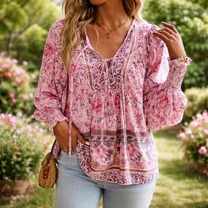 May include: A pink floral print blouse with long sleeves and a V-neck. The blouse features a tie at the neckline and elastic cuffs. The floral pattern includes pink, brown, and green elements. The sleeves are long and flowy.