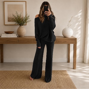 Könnte beinhalten: Ein schwarzes zweiteiliges Lounge-Set, bestehend aus einem langärmeligen Pullover mit schulterfreiem Ausschnitt und gerippten Bündchen sowie einer passenden Hose mit weitem Bein. Der Strickstoff weist eine feine vertikale Textur auf.