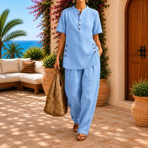 May include: A light blue short-sleeved shirt with a mandarin collar and a partial button-down front, paired with matching loose-fitting trousers. The shirt features decorative buttons along the side seams, and the outfit is worn with brown sandals.