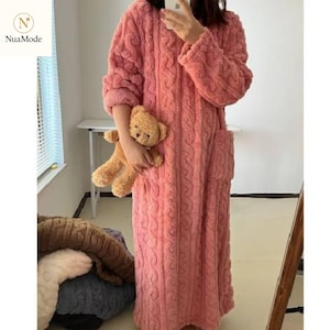 Pode incluir: Robe rosa comprido e macio com design de malha de cabo. O robe tem mangas compridas e dois bolsos. Um pequeno ursinho de pelúcia é segurado no bolso. O robe é feito de um material macio.