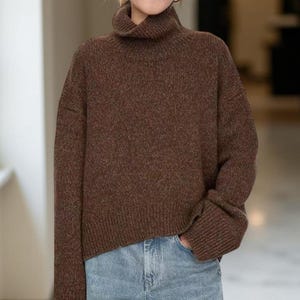 Può includere: Un maglione marrone a collo alto con maniche lunghe. Il maglione è realizzato in un tessuto a maglia strutturato e ha una vestibilità ampia. La persona indossa jeans blu.